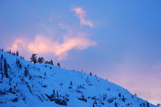 The sunset light hits a wispy cloud over a snow covered mountain