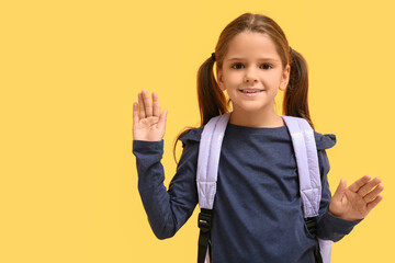Cute little happy girl with backpack waving hands on yellow background