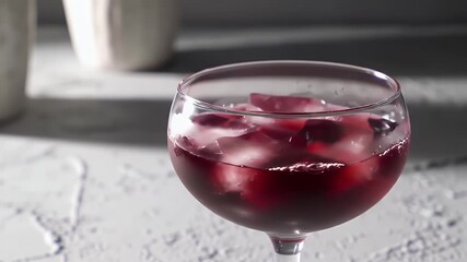 Crimson Beverage Cascading Into Stemmed Glass with Ice Cubes on Textured Light Gray Surface In High Angle Close Up Pouring Fresh Cold Cocktail For Restaurant Bar and Elegant Celebration