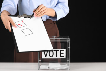 Voting businesswoman with clipboard near ballot box on black background