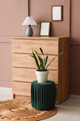 Obraz na płótnie Canvas Chest of drawers with lamp, blank frames and pouf near brown wall in room
