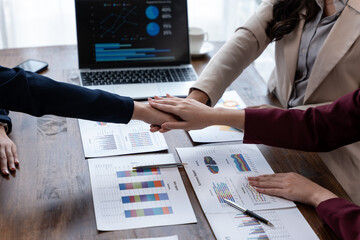 Team members stack hands over financial reports and charts on a desk, aligning on strategy during a...
