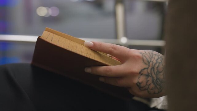 Close up of person with floral tattoo on hand flipping book page while reading indoors, showcasing natural nails and focus on hand movement with blurred indoor background