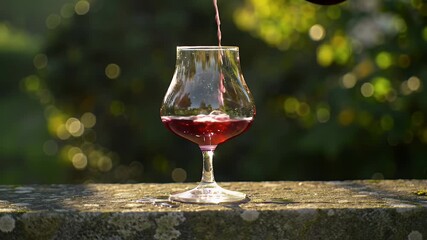Pouring Red Wine in Slow Motion into a Stemmed Glass on a Stone Ledge with Blurred Green Bokeh Background in Golden Light for Advertisement and Promotional Use in Wine Tasting Concepts
