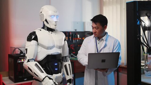 New York. April, 27 2020. Cybermetic robot employee cooperating with asian developer using laptop computer to control 3d printer automated machine. Additive manufacturing. AI.