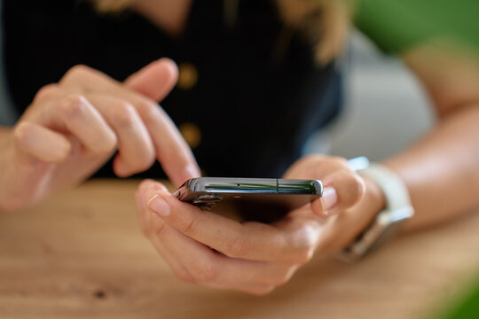 Close-up of woman sitting at table and using modern smartphone. Female hands holding mobile phone, browsing social media or messaging at home