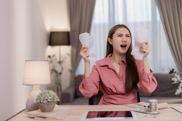 Young Asian woman tearing apart bills at her home office desk, showcasing frustration and stress stemming from financial difficulties and mounting economic pressures