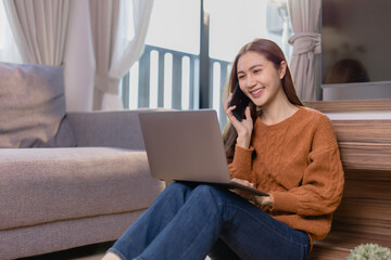 Naklejka premium Cheerful Asian freelancer working from home, sitting on the floor in her living room, using a laptop and talking on the phone, enjoying a comfortable and casual lifestyle