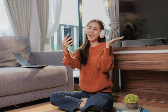 Happy young woman wearing headphones making a video call with her smartphone while sitting on the floor in her living room, with a laptop and tv in the background