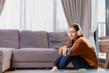 Relaxed young woman wearing headphones and holding a smartphone while listening to music, sitting on the floor in her living room, with a laptop on the sofa in the background