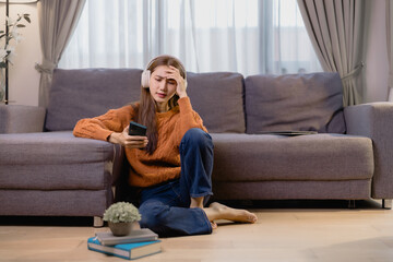 Young woman wearing headphones is sitting on the floor at home, holding her smartphone and suffering from headache, with a laptop on the sofa and books nearby