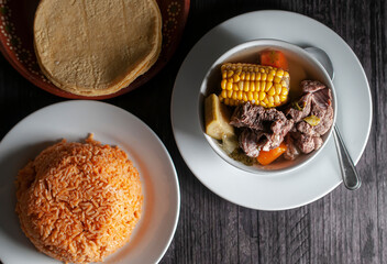 Beef rib broth with corn and carrots on a table with Mexican rice and corn tortillas, flat lay or top view