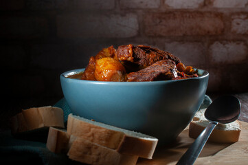 Beef ribs in spicy sauce inside a blue bowl with slices of white bread and a spoon on the sides, with a blurred brick wall in the background