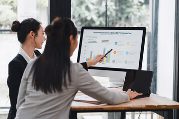 Young businesswoman presenting quarterly growth chart on desktop monitor to colleague in modern office, collaborative meeting with tablet and documents, focused professional discussion
