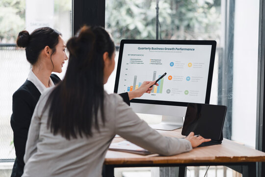 Young businesswoman presenting quarterly growth chart on desktop monitor to colleague in modern office, collaborative meeting with tablet and documents, focused professional discussion