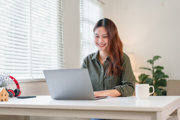 Fototapeta premium Young woman sitting at laptop smiling in bright home office with coffee and plant