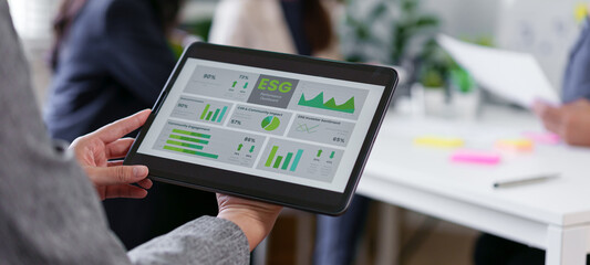 ESG performance dashboard on tablet displaying green charts and graphs, business team reviewing sustainability metrics in modern office, collaborative meeting with digital report