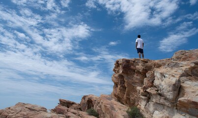 a man stands on top of the rocky cliff, looking down at us from above, with his back to the viewer. the sky is blue and clear, and there are some white clouds in it