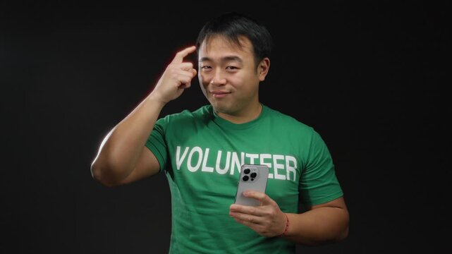 Young man with smartphone and green volunteer shirt poses confidently against black background emphasizing thoughtful expression.
