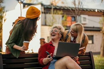 University students laughing together while using laptop on bench