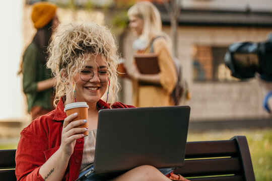 Smiling student working on laptop and drinking coffee outdoors on campus