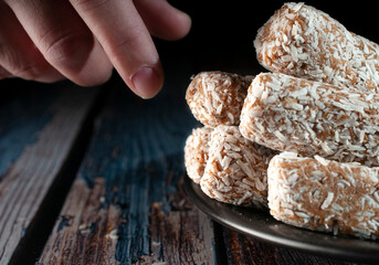 Hand taking sweet date rolls from a black plate on a rustic wooden table with a dark background. Macro photography of refined sugar and carbohydrate. horizontal image consumption.