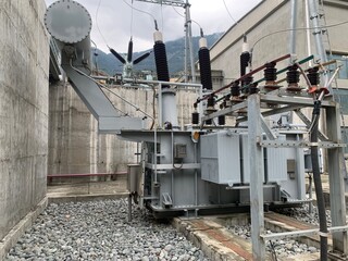 A large, high-voltage electrical transformer stands at an outdoor substation. The industrial equipment is set against a backdrop of a concrete wall and a rugged, mountainous landscape.