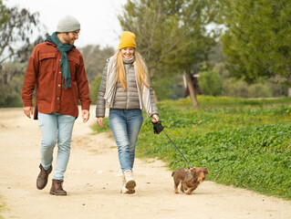 Smiling young couple walking their dachshund dog in a park, enjoying a leisurely stroll on a winter day ith copy space