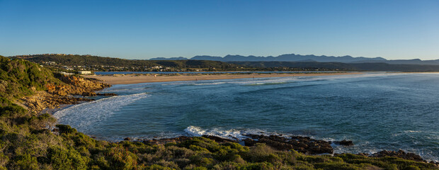 A breathtaking coastal view of Lookout Beach at Plettenberg Bay, Western Cape, South Africa, featuring dramatic waves, golden sand, lush greenery, and moody skies at sunset.