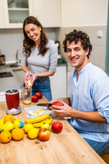 Adult couple is having fun while preparing smoothie in their kitchen. They are cutting fruits and using electric blender.