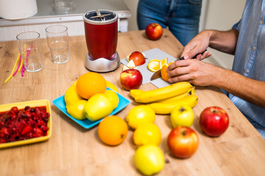 Close up of adult couple preparing smoothie in their kitchen. They are mixing various fruits in electric juicer. Man is slicing banana. - Powered by Adobe