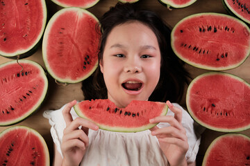 Portrait of a cheerful Asian young teenage girl eating many half pieces of fresh red watermelons,...