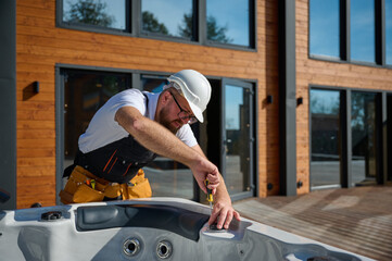 Plumber installing hot tub. Spa technician performing hot tub. Bathtub plumbing. Plumber fixing spa zone. Professional in uniform inspects and prepares a hot tub located on a stylish outdoor patio.