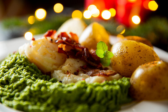 Traditional Christmas lutefisk dinner with pea puree, potatoes and bacon, served on porcelain plate with candles and spruce branches.