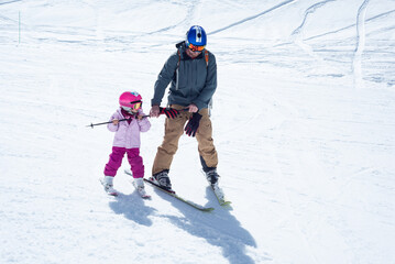A 3-year-old girl learning skiing with her father on a sunny winter day.