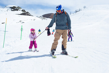 Front view of a father pulling his little daughter with his ski pole on a green slope during a nice ski day.
