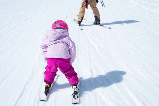 Rear view of a 3-year-old girl with a pink ski-wear skiing behaind her father.