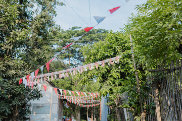 A festive outdoor scene in a rural neighborhood decorated with red and white triangular flags and tassels, strung across a small street. The flags, hanging between trees and bamboo fences