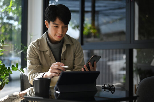 Young Asian man multitasking with a tablet and smartphone at a modern workspace, focused on digital tasks
