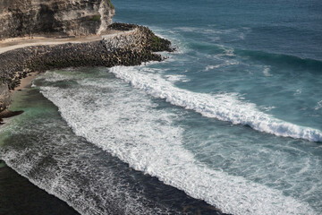 Waves crash on a rocky seawall at the base of a cliff in Bali, Indonesia. Scenic coastal view with powerful turquoise ocean surf.