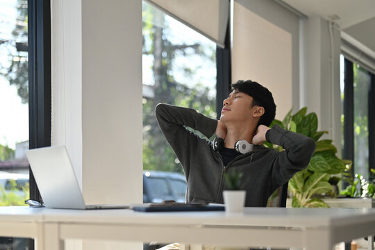Young man in hoodie leans back at his desk, stretching neck and relaxing after working on a laptop, enjoying a peaceful moment
