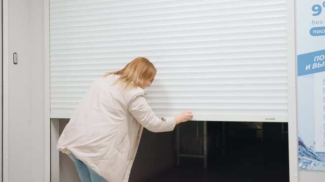 Laundry manager closes white rolling shutter at shop entrance, arms raised, dim interior with washing machine visible, corridor signage, casual jacket and jeans, clean indoor service scene