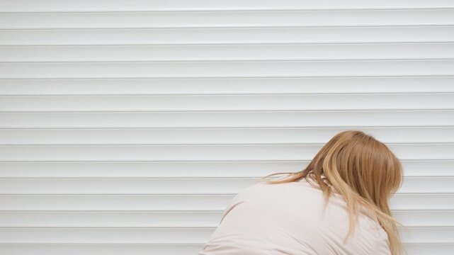 Back view of woman attendant rolling up white steel shutter to enter laundry room, hair down, light jacket, indoor setting, modern service facility with cart and washer faintly visible inside