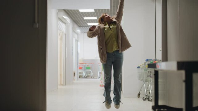 Laundry owner stands in corridor reaching upward as she jumps to pull down rolling steel door of laundry shop, wearing jacket and jeans, bright indoor setting with ceiling lights