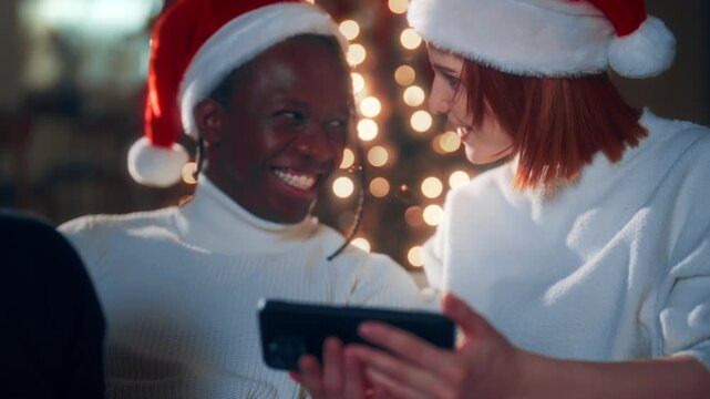 Wait for Christmas miracle, black man and Caucasian woman in Santa hats at home. Loving couple celebrating winter holidays together, using smartphone, watching funny video and pictures in internet