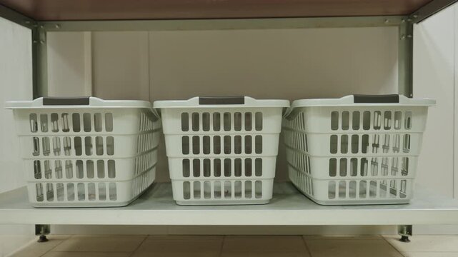 Three clothes hampers on metal shelf in tidy laundry room, empty plastic bins organized for service, clean utility storage scene with soft light, neutral tones, modern facility vibe