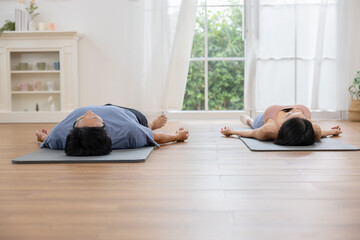 Asian Couple practicing yoga indoors lying on mats in bright room with wooden floor and large window, creating calm and peaceful atmosphere