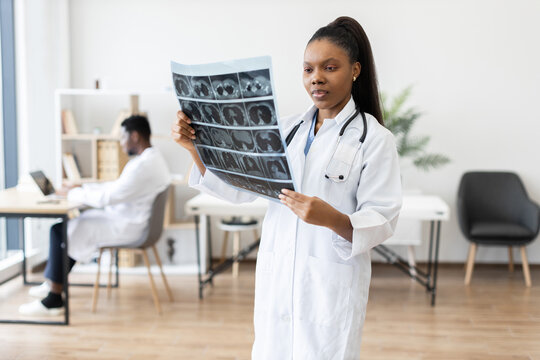 Female doctor analyzing X-ray scan in bright medical clinic. Background shows another doctor working. Highlights diagnostic evaluation, professional healthcare environment, teamwork.