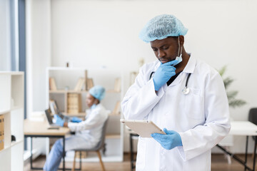 Male adult doctor wearing surgical attire and gloves reads medical information in clinic. Background nurse focuses on computer work, representing teamwork, medical professions