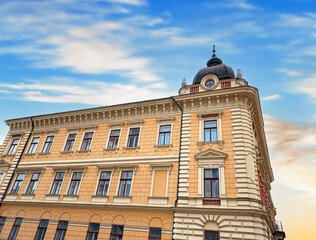 A historic European-style building with ornate architectural details, tall windows, and a domed corner roof stands against a bright sky in the city.